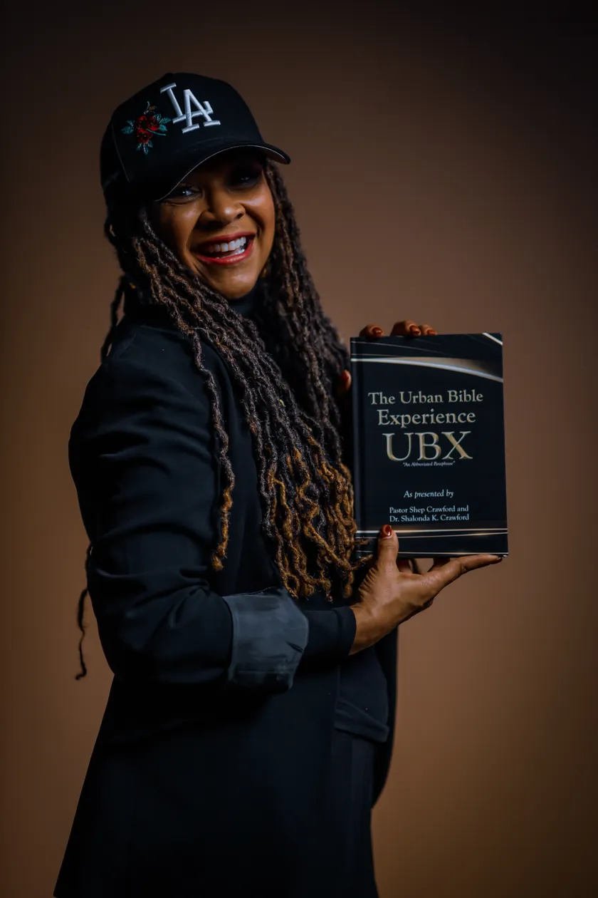 Dr. Shalonda &ldquo;Sunshine&rdquo; Crawford smiling and holding The Urban Bible Experience book, in a black LA cap and a black jacket