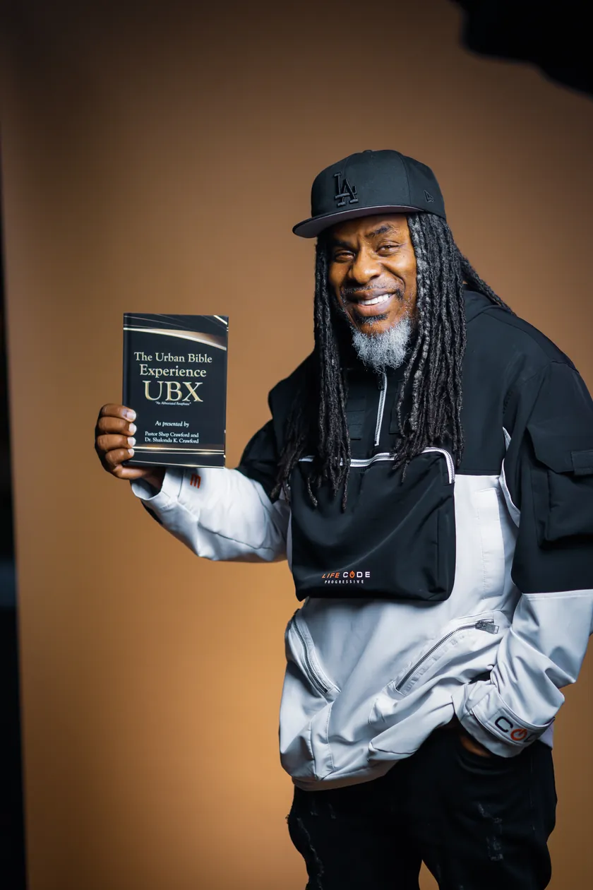 Pastor Shep Crawford smiling and holding up The Urban Bible Experience book, in a black LA cap and a black and white jacket